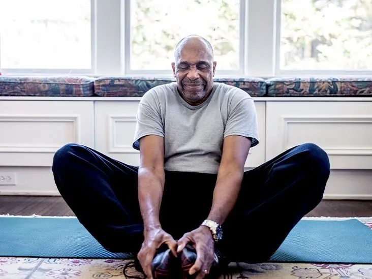 An older man sitting cross-legged on a yoga mat in his living room, eyes closed, gently stretching as part of a daily wellness routine